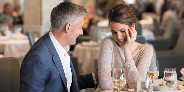 homem e mulher em roupas bonitas, sorrindo um para o outro, à mesa, com vinho branco