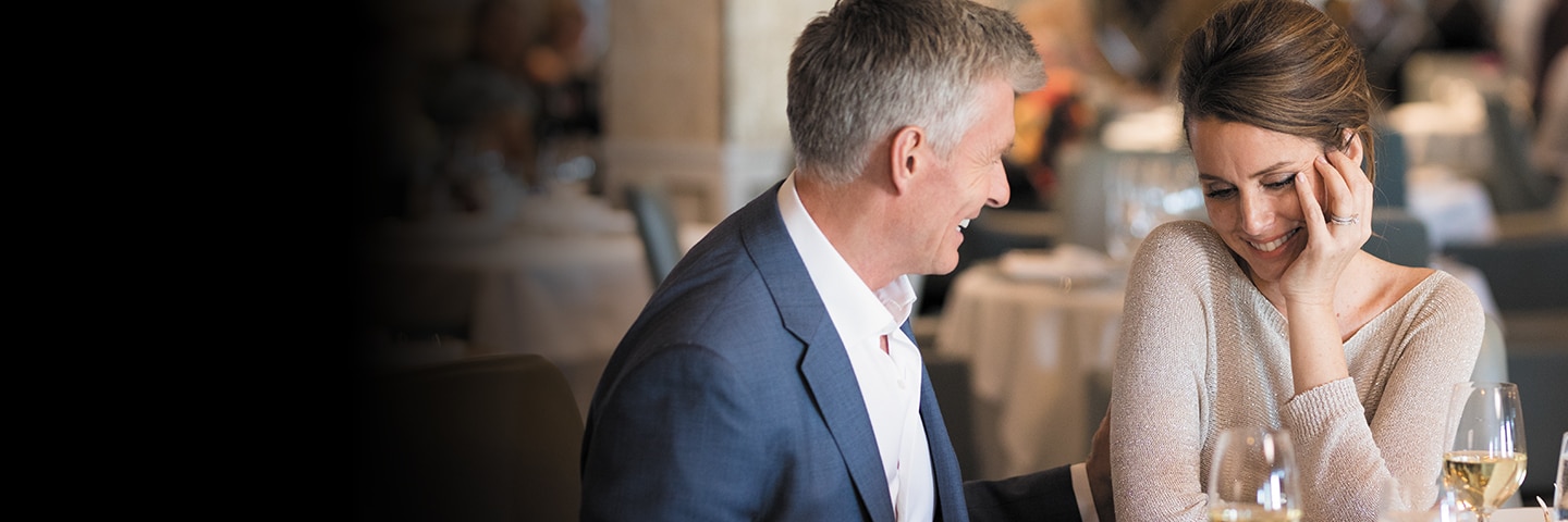 man and woman in nice clothes smiling at each other at table with white wine