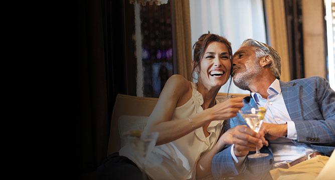 Ein Mann küsst eine lachende Frau mit einem Glas Martini in der Hand auf die Wange an Bord einer Neujahrs-Kreuzfahrt